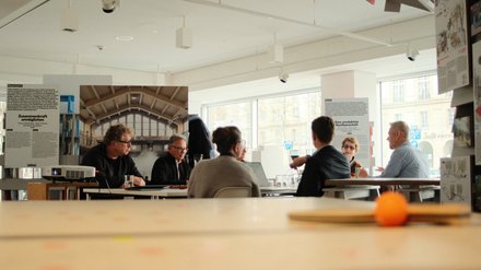 Mehrere Personen sitzen in einem hellen Raum an einem Tisch und diskutieren. Im Hintergrund sind Ausstellungswände und große Fenster zu sehen.