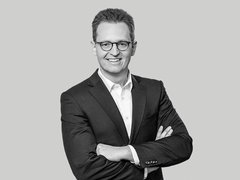 Schwarzweiß-Porträt eines Mannes im Anzug mit Brille, der mit verschränkten Armen in die Kamera lächelt.