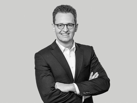 Schwarzweiß-Porträt eines Mannes im Anzug mit Brille, der mit verschränkten Armen in die Kamera lächelt.