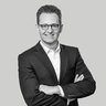 Schwarzweiß-Porträt eines Mannes im Anzug mit Brille, der mit verschränkten Armen in die Kamera lächelt.