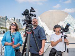 Drei Personen stehen auf einer Terrasse mit Filmausrüstung. Links eine Frau in hellblauem Hemd mit Sonnenbrille und buntem Halsschmuck hält ein Mikrofon. In der Mitte ein Mann in dunklem Poloshirt bedient eine große Filmkamera auf Stativ mit zusätzlichen Mikrofonen. Rechts ein Mann in weißem T-Shirt und schwarzer Kappe hält ein tragbares Aufnahmegerät. Im Hintergrund moderne Architektur mit weißem Kuppeldach, grüne Hochhäuser und blauer Himmel mit weißen Wolken."