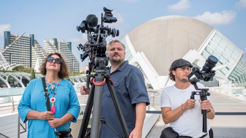 Drei Personen stehen auf einer Terrasse mit Filmausrüstung. Links eine Frau in hellblauem Hemd mit Sonnenbrille und buntem Halsschmuck hält ein Mikrofon. In der Mitte ein Mann in dunklem Poloshirt bedient eine große Filmkamera auf Stativ mit zusätzlichen Mikrofonen. Rechts ein Mann in weißem T-Shirt und schwarzer Kappe hält ein tragbares Aufnahmegerät. Im Hintergrund moderne Architektur mit weißem Kuppeldach, grüne Hochhäuser und blauer Himmel mit weißen Wolken."