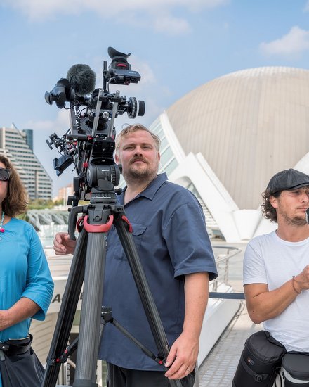 Drei Personen stehen auf einer Terrasse mit Filmausrüstung. Links eine Frau in hellblauem Hemd mit Sonnenbrille und buntem Halsschmuck hält ein Mikrofon. In der Mitte ein Mann in dunklem Poloshirt bedient eine große Filmkamera auf Stativ mit zusätzlichen Mikrofonen. Rechts ein Mann in weißem T-Shirt und schwarzer Kappe hält ein tragbares Aufnahmegerät. Im Hintergrund moderne Architektur mit weißem Kuppeldach, grüne Hochhäuser und blauer Himmel mit weißen Wolken."