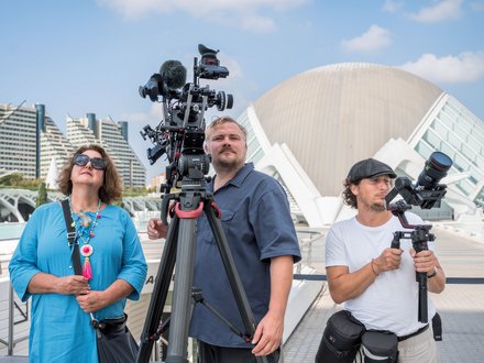 Drei Personen stehen auf einer Terrasse mit Filmausrüstung. Links eine Frau in hellblauem Hemd mit Sonnenbrille und buntem Halsschmuck hält ein Mikrofon. In der Mitte ein Mann in dunklem Poloshirt bedient eine große Filmkamera auf Stativ mit zusätzlichen Mikrofonen. Rechts ein Mann in weißem T-Shirt und schwarzer Kappe hält ein tragbares Aufnahmegerät. Im Hintergrund moderne Architektur mit weißem Kuppeldach, grüne Hochhäuser und blauer Himmel mit weißen Wolken."