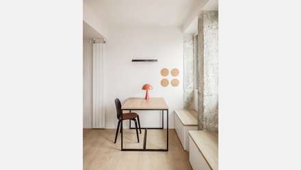 Innenraum mit einem Holztisch und einem schwarzem Stuhl vor einer weißen Wand; eine orangene pilzförmige Tischlampe steht auf dem Tisch 