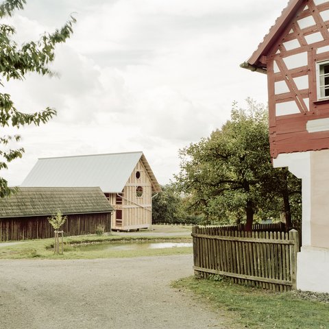 Eine Weitwinkelaufnahme einer ländlichen Szene im Freilichtmuseum. Im Vordergrund teilt sich ein Schotterweg. Links steht eine dunkle Holzscheune mit einem verwitterten Holzdach. Rechts sieht man die Ecke eines traditionellen Fachwerkhauses mit rotem Fachwerk und weißen Gefachen. Dahinter, in der Mitte, befindet sich ein neues, im Bau befindliches Gebäude in Holzrahmenbauweise mit einem glänzenden, gewellten Metalldach. Es hat eine runde und eine achteckige Fensteröffnung. Ein kleiner Teich liegt vor dem Neubau.