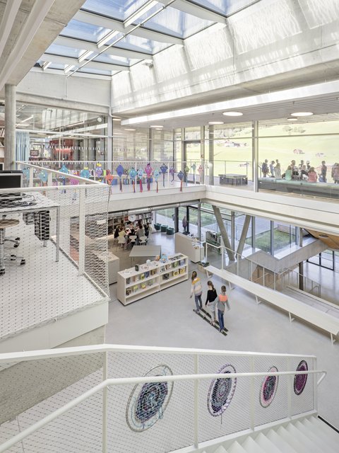 Blick über mehrere Ebenen eines lichtdurchfluteten Schulatriums mit Oberlichtern, Treppen und Netzgeländern. Kinder gehen durch die Bibliotheksbereiche; durch die Fenster ist eine grüne Landschaft sichtbar.