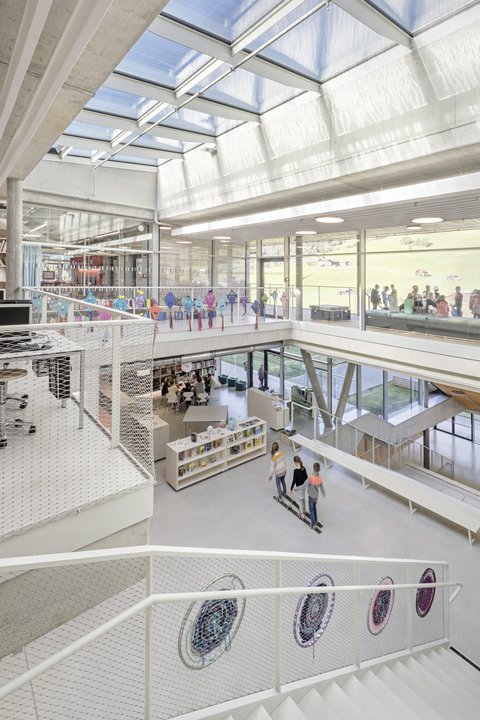 Blick über mehrere Ebenen eines lichtdurchfluteten Schulatriums mit Oberlichtern, Treppen und Netzgeländern. Kinder gehen durch die Bibliotheksbereiche; durch die Fenster ist eine grüne Landschaft sichtbar.