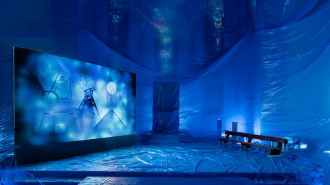 Blau beleuchteter Kirchenraum mit großformatiger Videoprojektion, verhüllten Flächen und Sitzbank als Teil einer künstlerischen Installation.