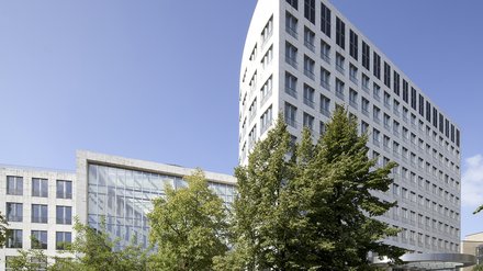 "Modernes Gebäudeensemble mit Wohn- und Geschäftshäusern. Weißes und graubetontes Hochhaus rechts mit regelmäßigen Fensterreihen. Niedrigeres weißes Gebäude mit großflächig verglaster Fassade links. Grüne Bäume im Vordergrund, gepflasterter öffentlicher Platz. Blauer Himmel."