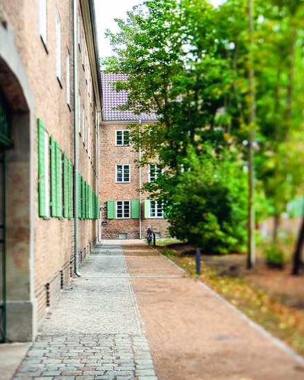 Backsteingebäude mit grünen Fensterläden entlang eines schmalen Weges, flankiert von Bäumen und Grünflächen.