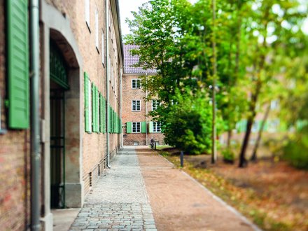 Backsteingebäude mit grünen Fensterläden entlang eines schmalen Weges, flankiert von Bäumen und Grünflächen.
