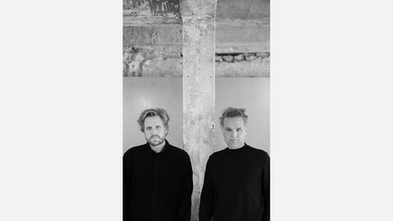 Zwei Männer stehen nebeneinander vor einer Betonstütze in einem unfertigen Innenraum.