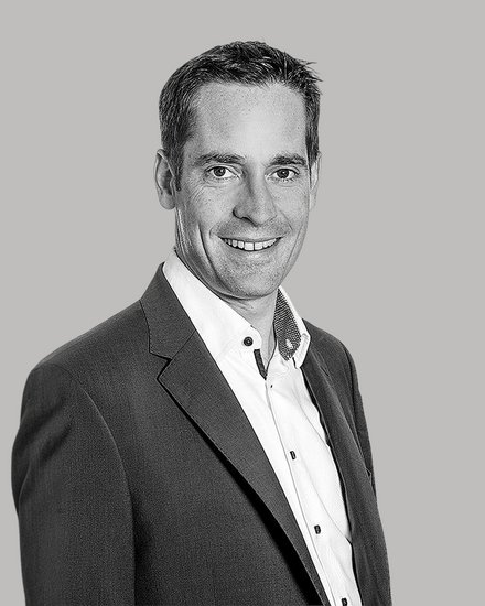 Schwarzweiß-Porträt eines lächelnden Mannes im Anzug und weißen Hemd, leicht seitlich zur Kamera stehend vor neutralem Hintergrund.