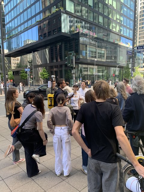 Eine Menschengruppe steht auf einem städtischen Gehweg vor modernen Bürogebäuden und hört einer Person mit einem gelben Koffer-Rucksack zu.