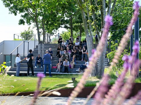 "Breite dunkle Betontreppen in einem Park, auf denen etwa 30-40 Personen sitzen und stehen. Ein Mann und eine Frau im schwarzen Outfit moderieren die Veranstaltung unten links. Die Treppen sind umgeben von hohen grünen Laubbäumen und blühenden Stauden mit lila-rosa Blüten (Phlox) im Vordergrund. Klarer blauer Himmel mit leichten Wolken. Moderne Architektur und Betonelemente rahmen den Raum ein."