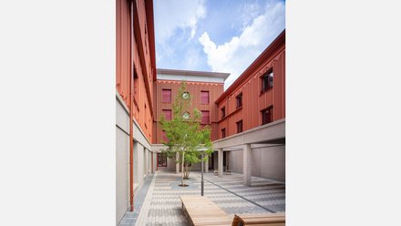 Innenhof mit roten Fassaden, Betonarkaden und Sitzbänken; mittig ein Baum, blauer Himmel mit Wolken.