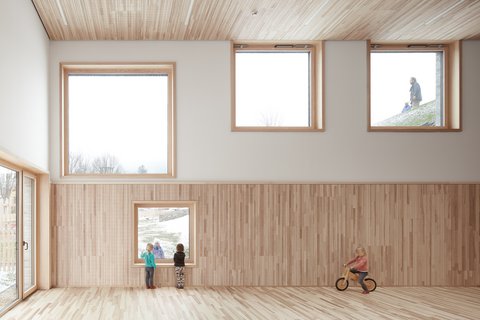 Heller Raum mit Holzverkleidung an der Wand, mehreren quadratischen Fenstern und einem Kind auf einem Laufrad