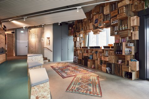 Innenraum mit Sitzgelegenheiten, Teppichen und einer wandfüllenden Regalinstallation aus gestapelten Holzkisten