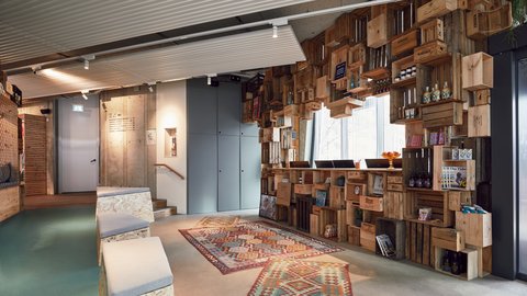 Innenraum mit Sitzgelegenheiten, Teppichen und einer wandfüllenden Regalinstallation aus gestapelten Holzkisten