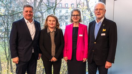 Vier erwachsene Personen stehen nebeneinander vor einer großen Fensterfront; sie tragen formelle Kleidung und blicken in die Kamera, im Hintergrund sind Bäume zu sehen.
