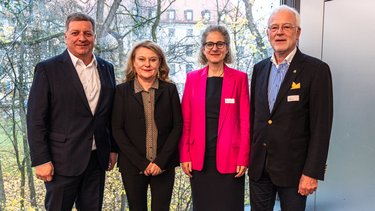 Vier erwachsene Personen stehen nebeneinander vor einer großen Fensterfront; sie tragen formelle Kleidung und blicken in die Kamera, im Hintergrund sind Bäume zu sehen.