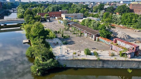 Eine Drohnenaufnahme eines urbanen Geländes an einem Flusslauf. Zu sehen sind Container mit Graffiti, eine offene Halle, viel Grün und im Hintergrund die Silhouette einer Stadt.