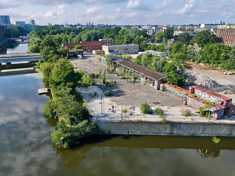 Eine Drohnenaufnahme eines urbanen Geländes an einem Flusslauf. Zu sehen sind Container mit Graffiti, eine offene Halle, viel Grün und im Hintergrund die Silhouette einer Stadt.