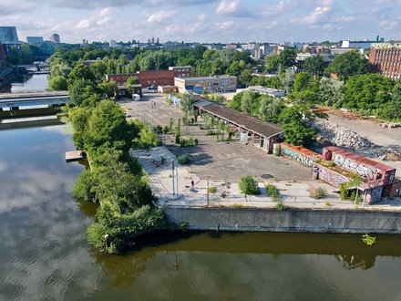 Eine Drohnenaufnahme eines urbanen Geländes an einem Flusslauf. Zu sehen sind Container mit Graffiti, eine offene Halle, viel Grün und im Hintergrund die Silhouette einer Stadt.