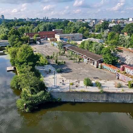 Eine Drohnenaufnahme eines urbanen Geländes an einem Flusslauf. Zu sehen sind Container mit Graffiti, eine offene Halle, viel Grün und im Hintergrund die Silhouette einer Stadt.