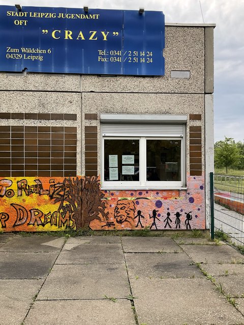  Nahaufnahme der Fassade eines Betongebäudes des "Stadt Leipzig Jugendamt OFT 'Crazy'". Ein großes blaues Schild oben zeigt den Namen und die Adresse. Darunter befindet sich ein Fenster mit Jalousien und Zetteln. Der Sockel ist bunt mit Graffiti bemalt, darunter der Schriftzug "TO REALIZE YOUR DREAMS", ein Tierkopf und Strichmännchen. Ein Metallzaun ist rechts zu sehen.