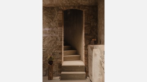 "Steingewölbe mit rustikalen Backsteinmauern. Eine dunkle Holztür mit Rahmen wird von zwei breiten Stufen erreicht. Links neben der Tür ein kleiner Topf mit Pflanze auf einem Holzständer. Der Durchgang ist eng und führt in Dunkelheit. Warmes Steinambiente."