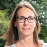 Porträt einer Person mit Brille und schulterlangem Haar im Freien; gestreiftes T‑Shirt, Blick in die Kamera.