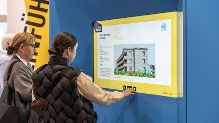 Zwei Besucherinnen an einer interaktiven Infostation: eine jüngere Frau tippt auf den großen Touchscreen, eine ältere Frau schaut zu; Ausstellungsszenen und gelbe Gestaltungselemente sichtbar.