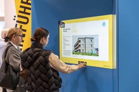 Zwei Besucherinnen an einer interaktiven Infostation: eine jüngere Frau tippt auf den großen Touchscreen, eine ältere Frau schaut zu; Ausstellungsszenen und gelbe Gestaltungselemente sichtbar.