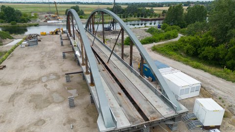 Luftaufnahme: Eine im Bau befindliche Stahlbogenbrücke steht auf provisorischen Stützen am Fluss. Das Fahrbahndeck ist noch offen; nebenan stehen Baucontainer, Bagger und Kräne. Im Hintergrund liegen Felder, Baumreihen und mehrere Windräder.