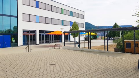 Weiße Schulgebäude mit bunten Sonnenschirmen und überdachtem Außenbereich vor bewaldeten Hügeln.