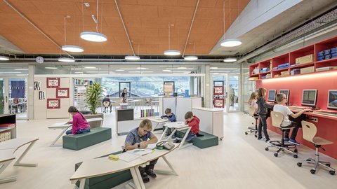 Lern- und Bibliotheksbereich unter Oberlichtern: Schülerinnen und Schüler arbeiten an Tischen, nutzen Computer und lesen gemeinsam an einem Regal. Durch die Fenster sind Berge zu sehen.