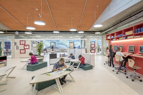 Lern- und Bibliotheksbereich unter Oberlichtern: Schülerinnen und Schüler arbeiten an Tischen, nutzen Computer und lesen gemeinsam an einem Regal. Durch die Fenster sind Berge zu sehen.