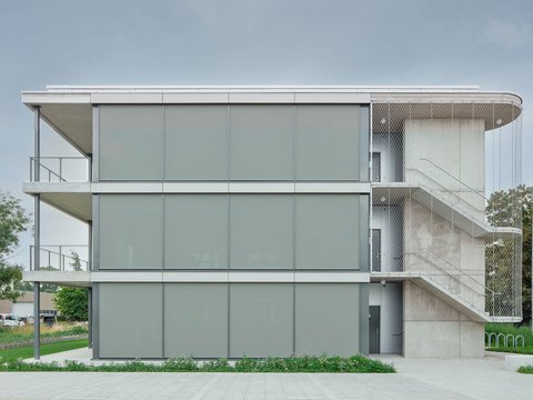 „Dreigeschossiges modernes Gebäude mit grauer Fassade und umlaufenden Balkonen. Rechts führt eine offene Betontreppe hinter einem Metallnetz zu den oberen Etagen.“