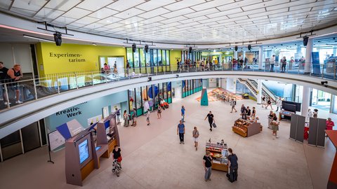 Innenansicht eines zweigeschossigen Gebäudes mit Besuchern, gelben und blauen Wänden mit den Schriftzügen 'Experimentarium' und 'Kreativ-Werk'