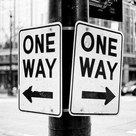 Schwarz‑weiß‑Foto: Zwei an einem Laternenmast befestigte Verkehrsschilder mit der Aufschrift „ONE WAY“, eins mit Pfeil nach links, das andere mit Pfeil nach rechts; unscharfer Stadthintergrund mit Gebäuden, Ampeln und Fahrzeugen.