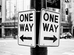 Schwarz‑weiß‑Foto: Zwei an einem Laternenmast befestigte Verkehrsschilder mit der Aufschrift „ONE WAY“, eins mit Pfeil nach links, das andere mit Pfeil nach rechts; unscharfer Stadthintergrund mit Gebäuden, Ampeln und Fahrzeugen.