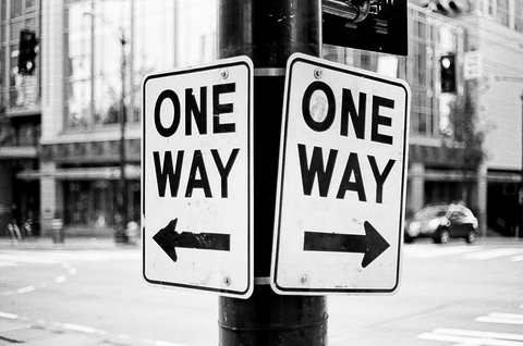 Schwarz‑weiß‑Foto: Zwei an einem Laternenmast befestigte Verkehrsschilder mit der Aufschrift „ONE WAY“, eins mit Pfeil nach links, das andere mit Pfeil nach rechts; unscharfer Stadthintergrund mit Gebäuden, Ampeln und Fahrzeugen.