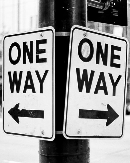 Schwarz‑weiß‑Foto: Zwei an einem Laternenmast befestigte Verkehrsschilder mit der Aufschrift „ONE WAY“, eins mit Pfeil nach links, das andere mit Pfeil nach rechts; unscharfer Stadthintergrund mit Gebäuden, Ampeln und Fahrzeugen.