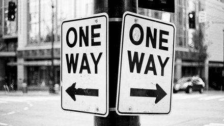 Schwarz‑weiß‑Foto: Zwei an einem Laternenmast befestigte Verkehrsschilder mit der Aufschrift „ONE WAY“, eins mit Pfeil nach links, das andere mit Pfeil nach rechts; unscharfer Stadthintergrund mit Gebäuden, Ampeln und Fahrzeugen.