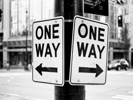 Schwarz‑weiß‑Foto: Zwei an einem Laternenmast befestigte Verkehrsschilder mit der Aufschrift „ONE WAY“, eins mit Pfeil nach links, das andere mit Pfeil nach rechts; unscharfer Stadthintergrund mit Gebäuden, Ampeln und Fahrzeugen.