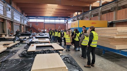 Werkhalle zur Holzbau‑Fertigung: Gruppe in Warnwesten auf Werksführung; Holz‑Wand/Deckenelemente auf Planen, gestapelte Bretter, Maschinen.