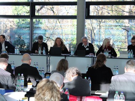 Mehrere Personen sitzen hinter einem langen Tisch vor großen Fenstern mit Blick ins Freie, arbeiten an Laptops oder hören zu, während davor weitere Teilnehmende Platz genommen haben, wodurch das Bild eine Gremiensitzung oder Fachdebatte dokumentiert und für strukturierte Zusammenarbeit und gemeinsame Entscheidungsfindung steht.
