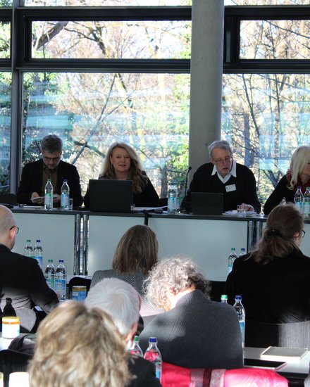 Mehrere Personen sitzen hinter einem langen Tisch vor großen Fenstern mit Blick ins Freie, arbeiten an Laptops oder hören zu, während davor weitere Teilnehmende Platz genommen haben, wodurch das Bild eine Gremiensitzung oder Fachdebatte dokumentiert und für strukturierte Zusammenarbeit und gemeinsame Entscheidungsfindung steht.